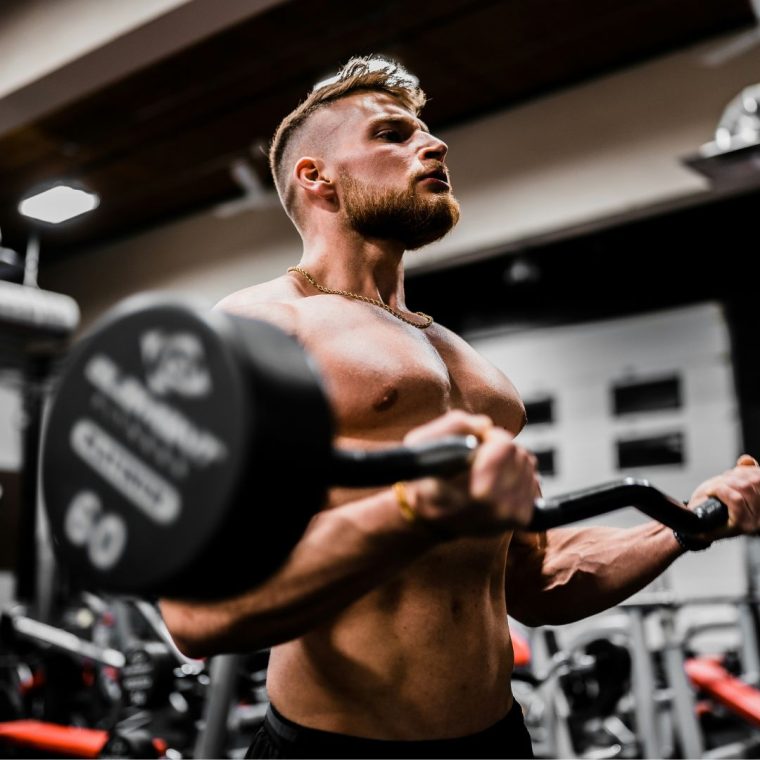A muscular man with a beard is lifting a heavy barbell inside a gym, reminiscent of the dedication required to enhance ROI in an ecommerce store. He is shirtless, wearing black shorts, and appears focused on his workout. The gym equipment and setting are visible in the background, with a slightly blurred effect.