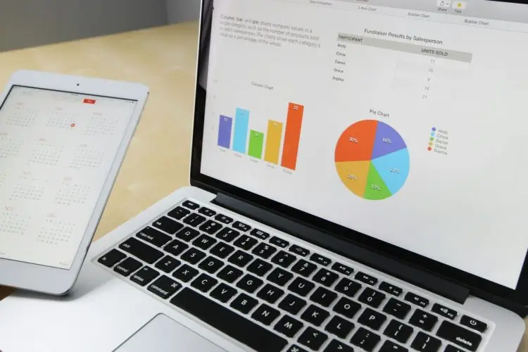 A person holds a tablet displaying a monthly calendar in front of a laptop screen. The laptop shows various charts and graphs, including a pie chart and bar graph, representing sales data and statistics. This scene is set on a light wooden desk, illustrating the analytical tools used by digital marketing agencies.