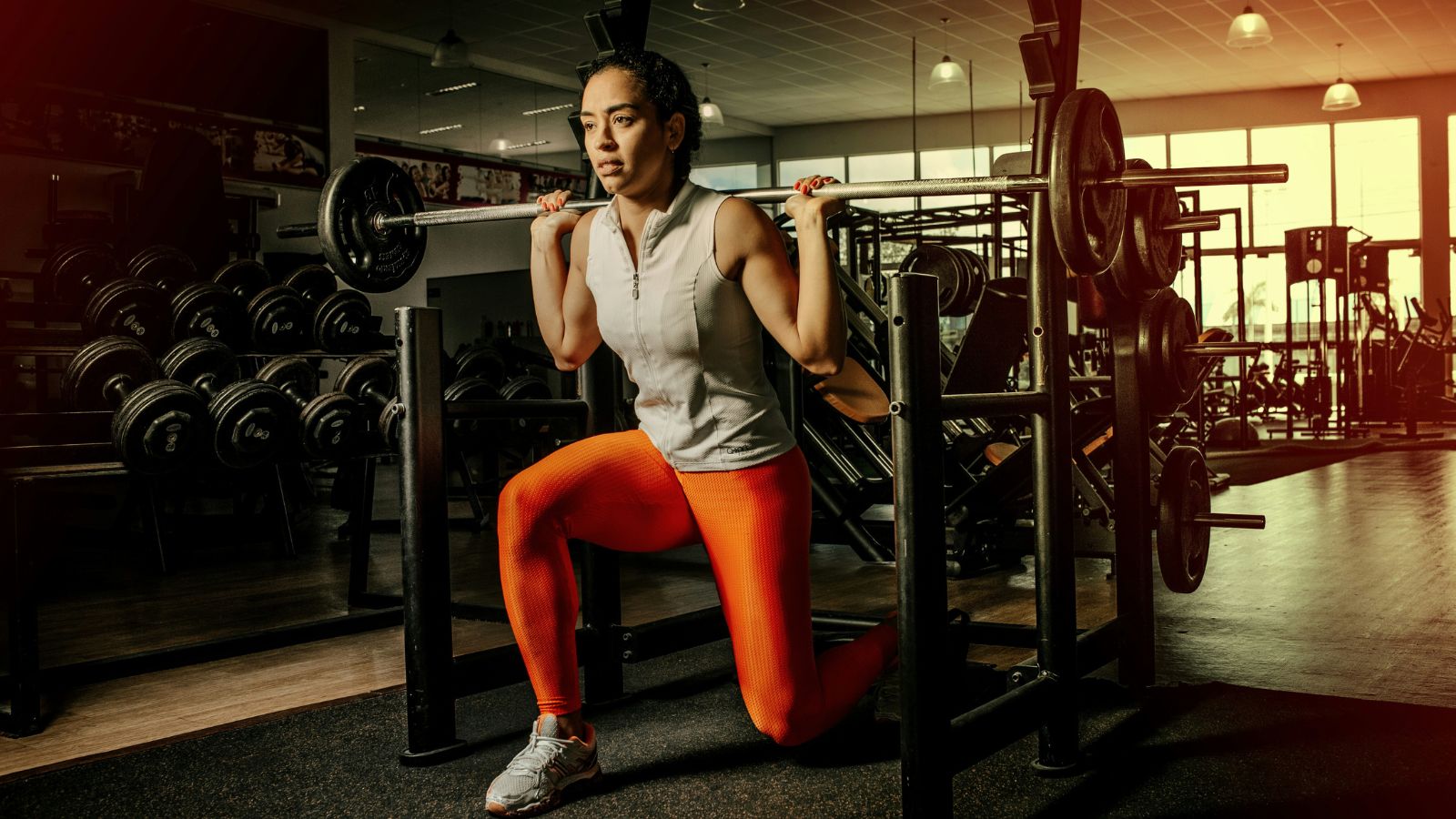 8x ROAS for a Fitness Center 17 A person wearing a sleeveless top and bright orange leggings performs a barbell squat in a gym. The gym, which is brightly lit with large windows, is equipped with various weightlifting machines, racks, and dumbbells. This well-equipped setting could enhance ROI for those promoting fitness equipment on Facebook ads for Achieving 8x ROAS for a Fitness Center through Google Ads